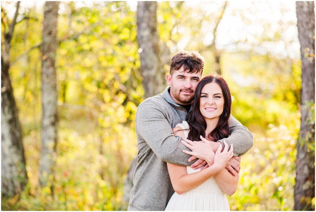 man wrapping his arms around the front of his fiance while standing in front of some trees 