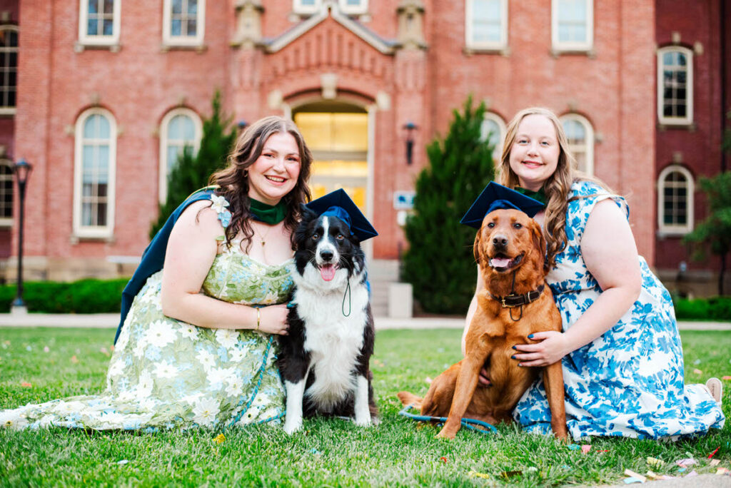 two smiling graduates in floral dresses sit on the grass with their dogs both wearing blue graduation caps in front of woodburn hall during wvu grad photos