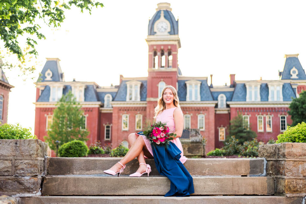wvugradphotos008 carrie white photography smiling woman in a pink dress and blue skirt sits on stone steps holding a pink bouquet with a clock tower campus building behind her during wvu spring grad photos