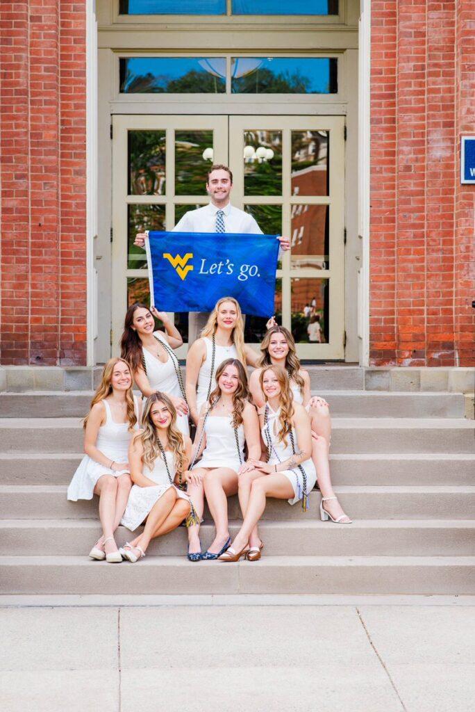 group of smiling women in white dresses posing on campus steps man behind them holding a blue banner that says lets go with a yellow wv logo