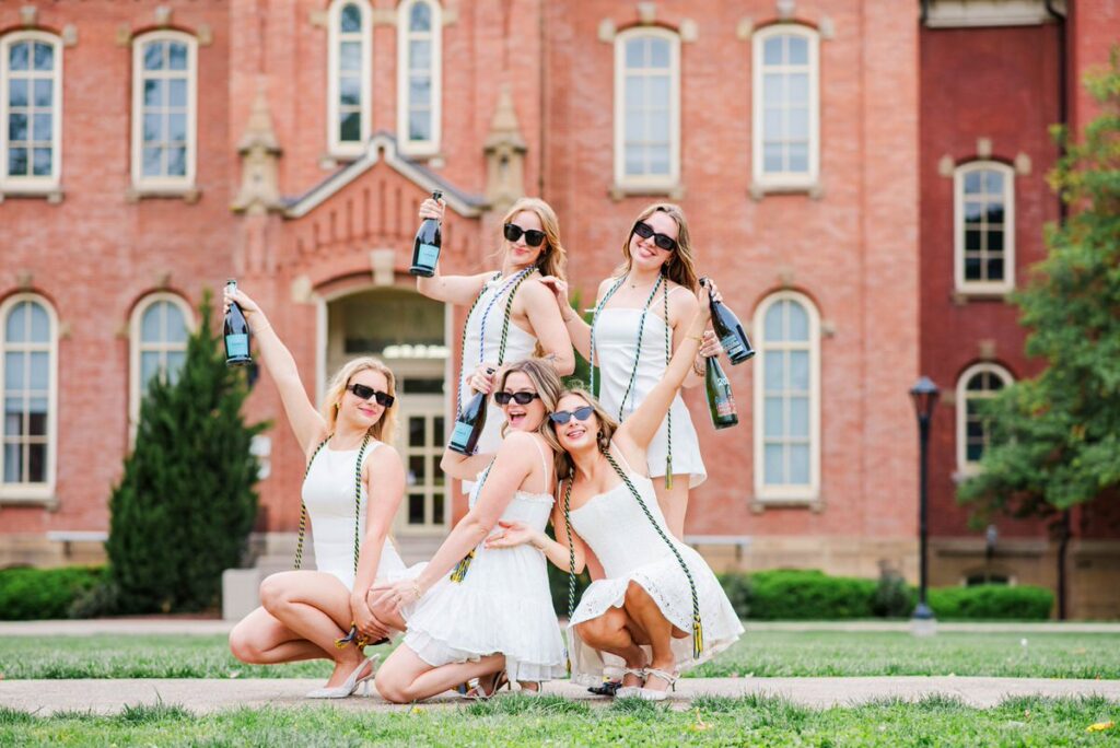 group of women in white dresses posing outdoors with champagne bottles in front of a brick university building celebrating together