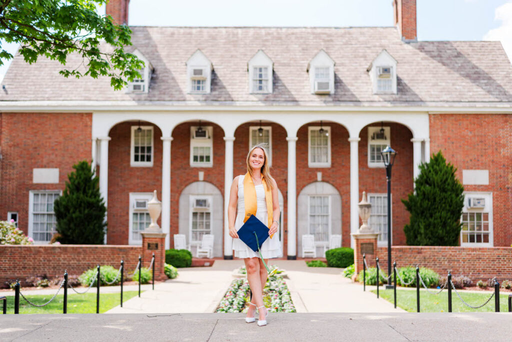 wvugradphotos019 carrie white photography female graduate in a white dress and orange stole stands on a campus path with a blue graduation cap posing in front of a brick building with arches
