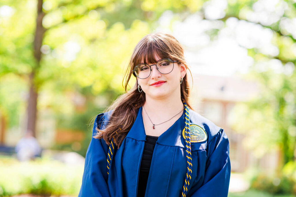 wvugradphotos021 carrie white photography young woman in a blue graduation gown and cords posing outdoors on a sunny campus smiling at the camera