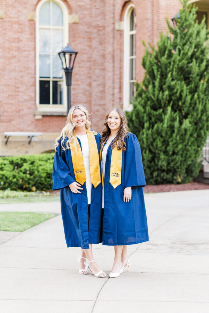 two graduates in blue gowns and gold stoles pose outside a brick campus building on a sunny day
