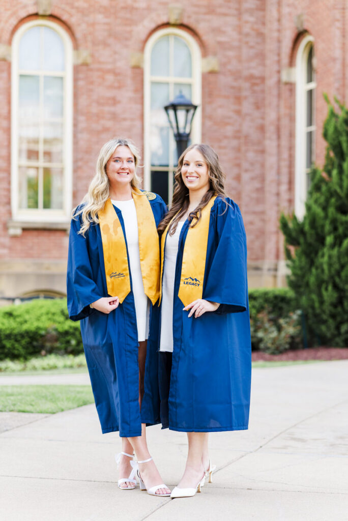 wvugradphotos027 carrie white photography two graduates in blue gowns with yellow stoles stand together outdoors smiling in front of a red brick campus building