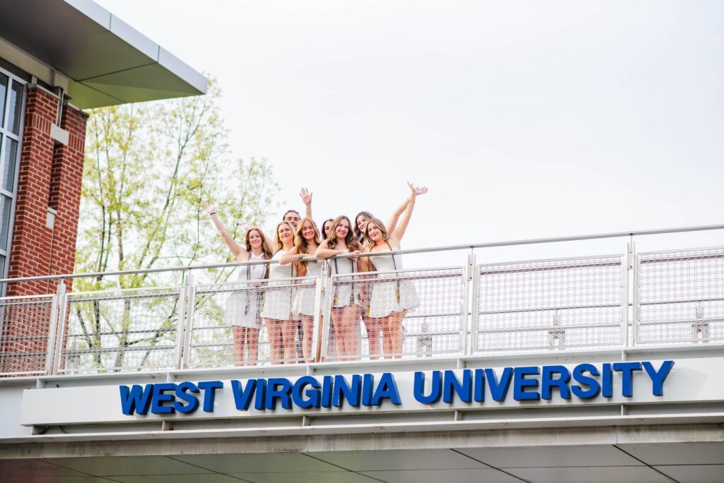 group of smiling students on a balcony above a sign reading'West Virginia University' with waving hands up in the air.