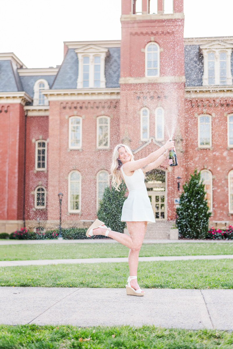 Woman in a white dress sprays champagne outdoors, one leg lifted, in front of a red brick campus building.
