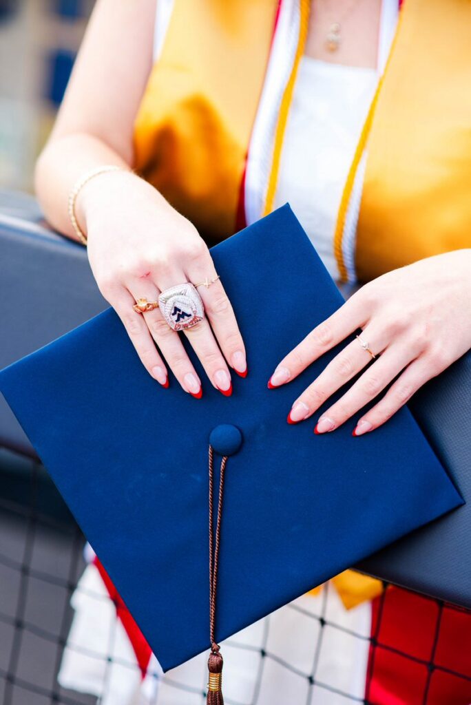wvugradphotos076 carrie white photography graduate's hands with red nails holding a blue graduation cap with tassel, wearing a yellow gown or stole in the background.