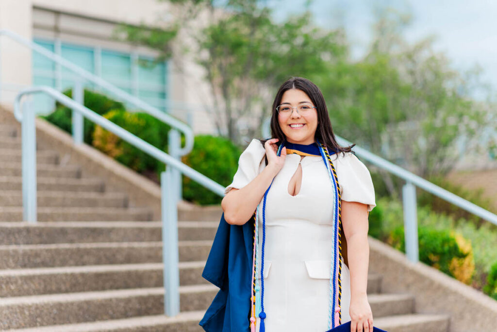 graduate in a white dress and blue cap and gown with cords standing on outdoor steps with a campus building in the background