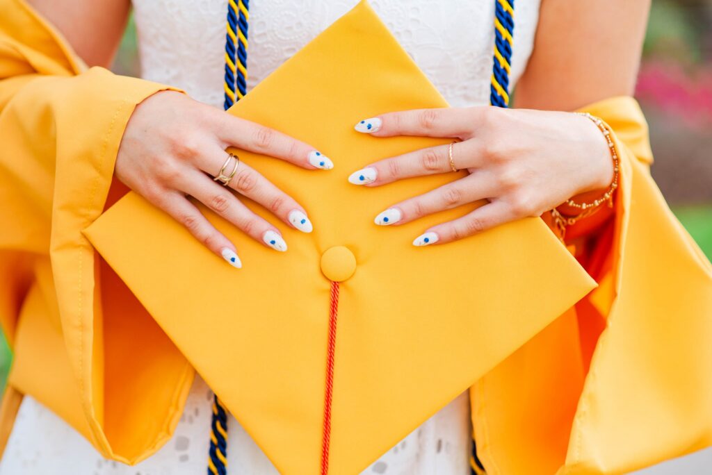 wvugradphotos11 carrie white photography person holding a yellow graduation cap with blue and yellow tassel wearing an orange gown and white dress underneath close up of hands with blue dotted nails and simple rings