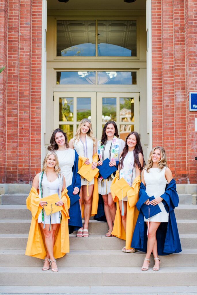 six young graduates stand on campus steps in white dresses and blue and yellow gowns holding matching caps outdoors in front of a brick building