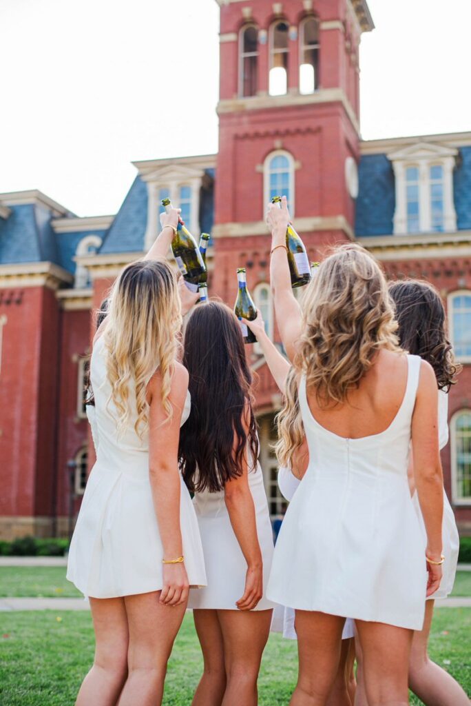 a group of women in white dresses raising champagne bottles in celebration outdoors in front of woodburn hall in morgantown wv
