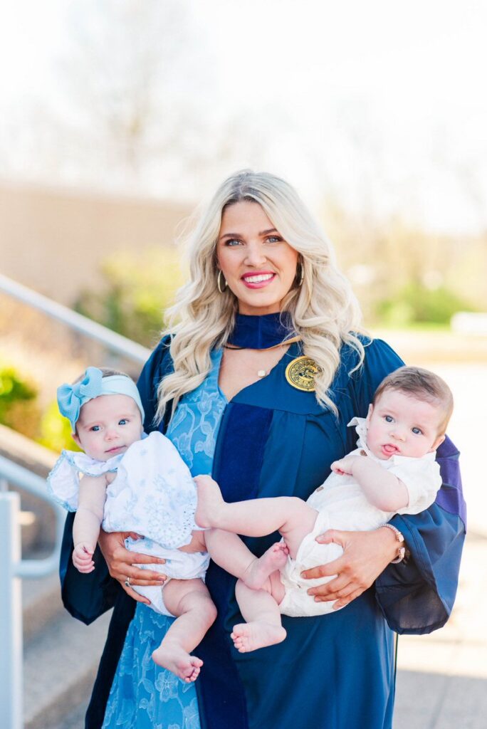 graduate mother in a navy blue gown holding two babies outdoors smiling at the camera