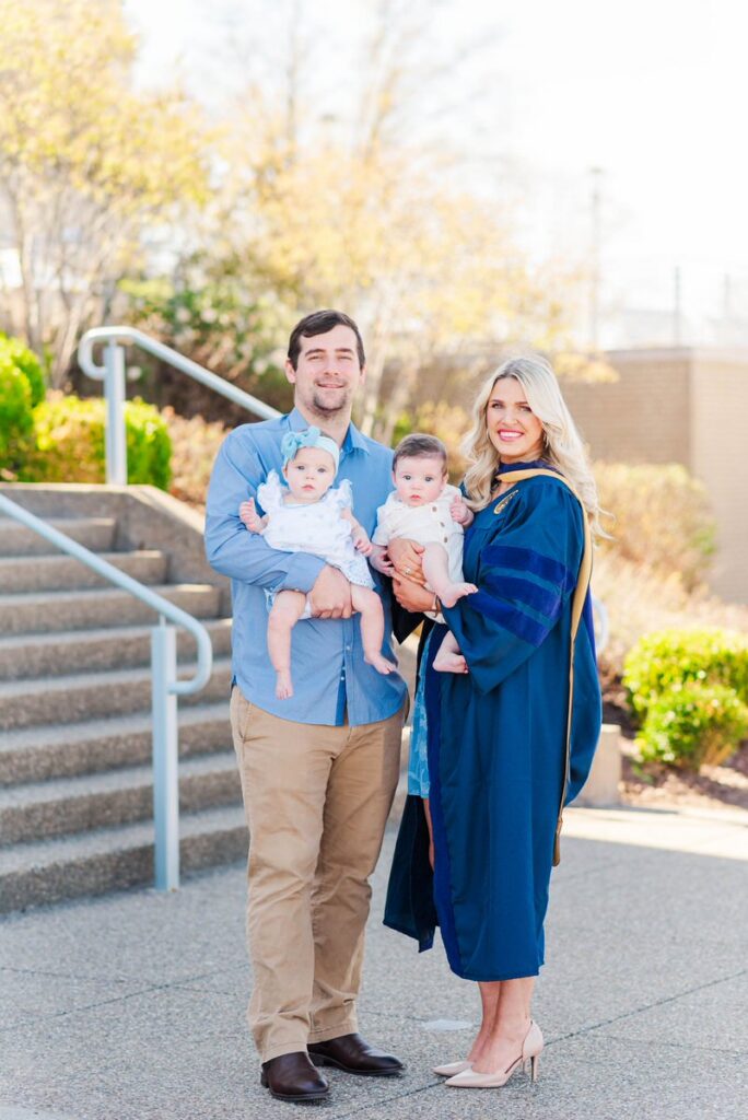 family portrait of a man and woman holding two babies outdoors the mother wears a blue graduation gown and beige heels smiling at the camera