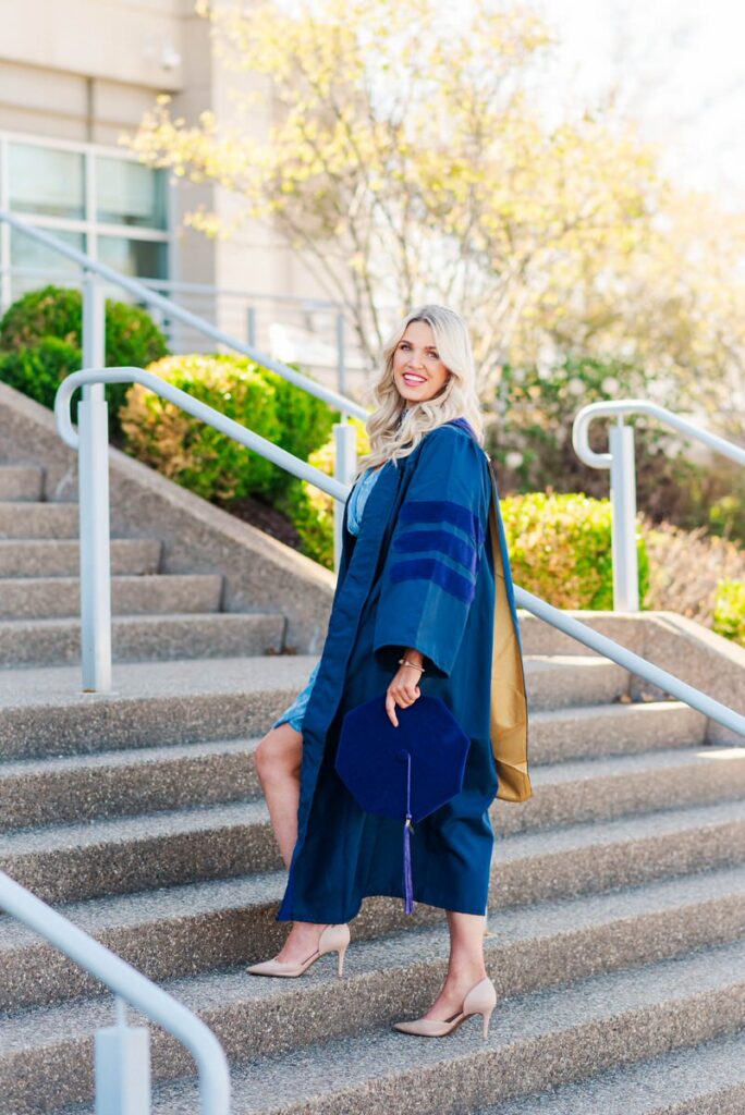 graduate in a navy blue gown and cap descends outdoor stairs smiling holding her cap with a purple tassel and carrying a tan tote bag