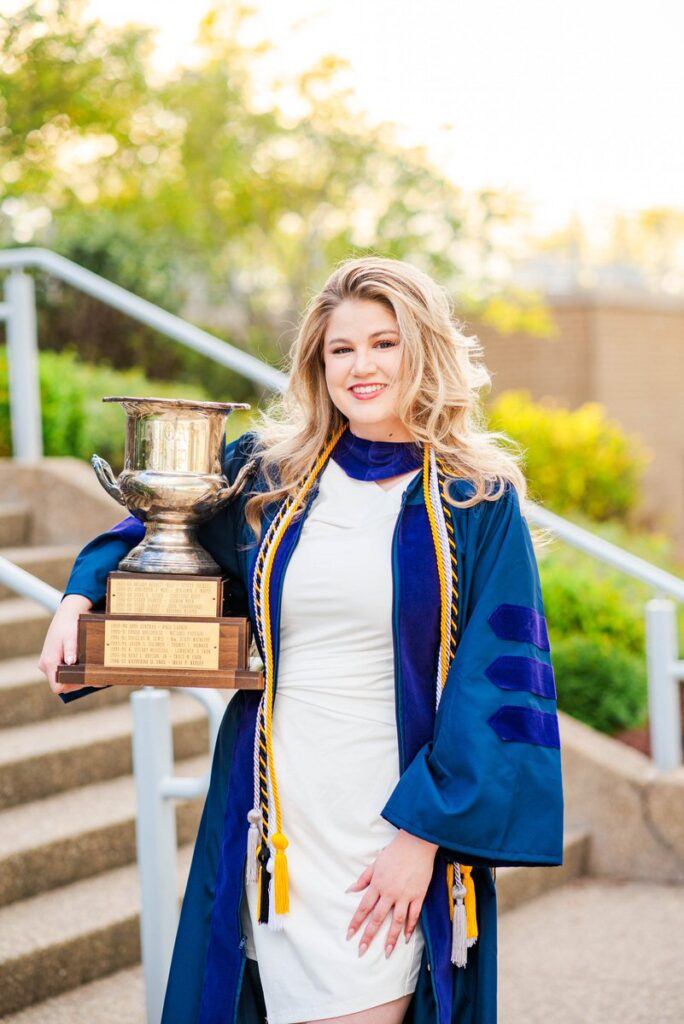 graduate in a blue robe smiles while holding a large trophy outdoors on campus