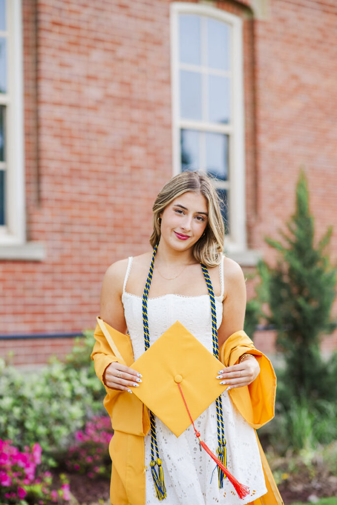 wvugradphotos151 carrie white photography young woman in a white dress wearing blue and gold graduation cords holding a yellow cap in front of a brick building