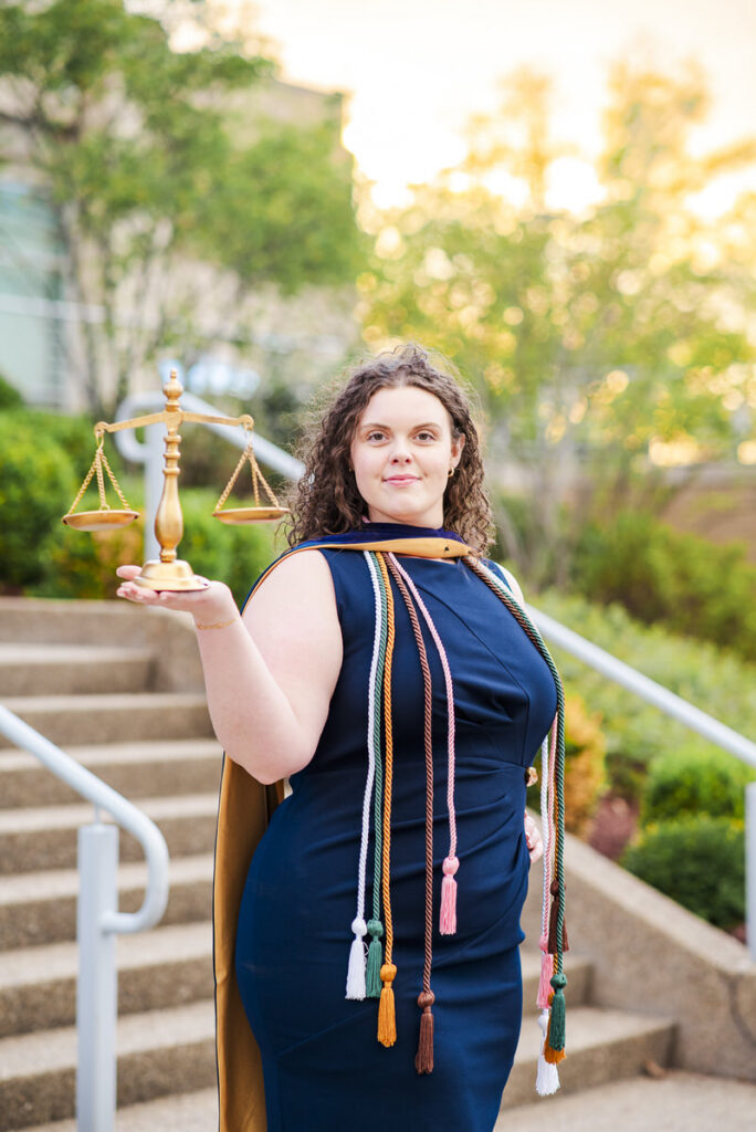 woman in a navy dress and cape holding a gold scales of justice wearing multiple ceremonial cords outdoors on steps