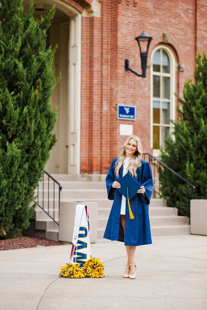 graduate in a blue cap and gown posing outside a brick university building holding her cap with a yellow tassel
