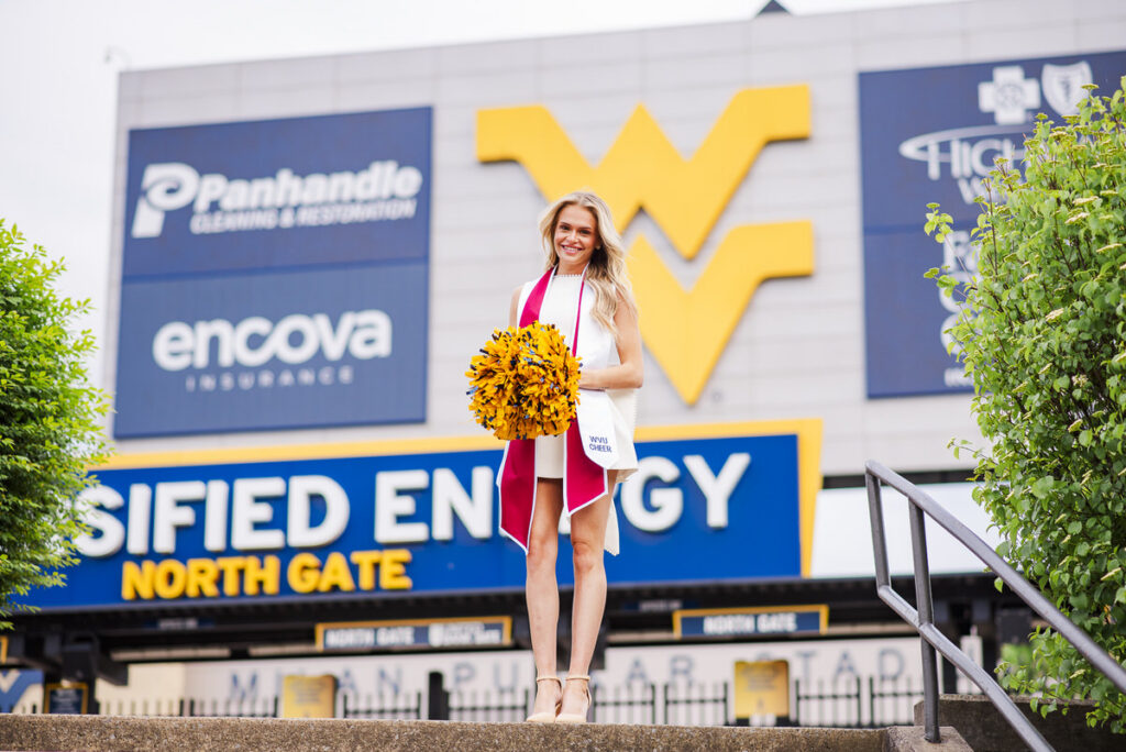 wvugradphotos187 carrie white photography cheerleader in white and pink with yellow pom poms posing on stadium steps in front of sponsor banners and a large wvu style logo