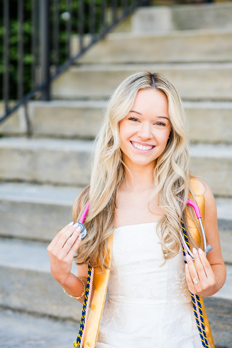 smiling young woman in a white dress with a pink stethoscope posing on outdoor steps after graduation wearing a yellow sash and cords