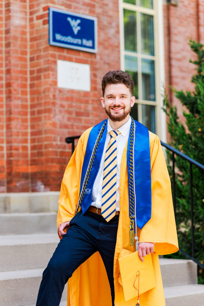 graduate in a gold gown with a blue stole holding a diploma envelope posing on campus stairs outside a brick building