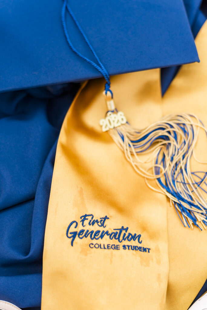 blue graduation cap with a gold sash reading'First Generation College Student' and a '2026' charm, symbolizing a first-generation graduate.