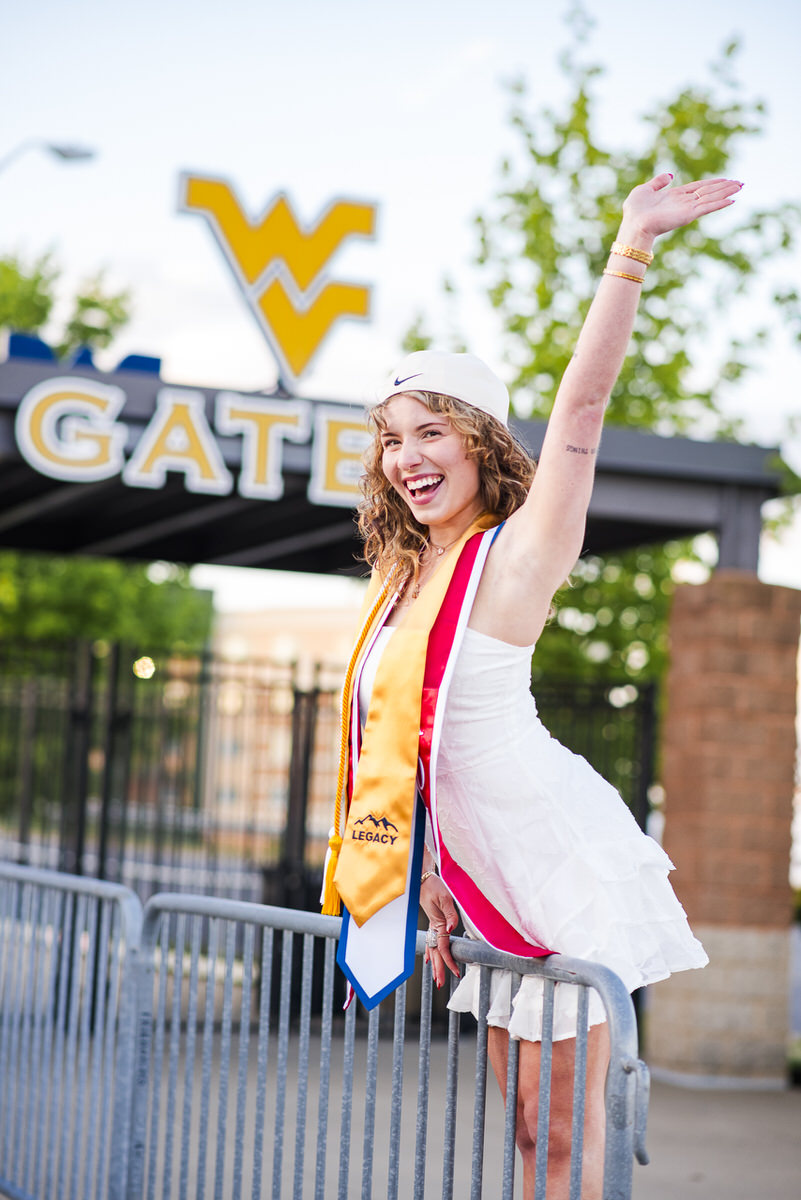 graduate in a white dress and cap raises her arm in celebration beside a metal gate and a wv signbanner she wears gold honor cords and a yellow ribbon sash