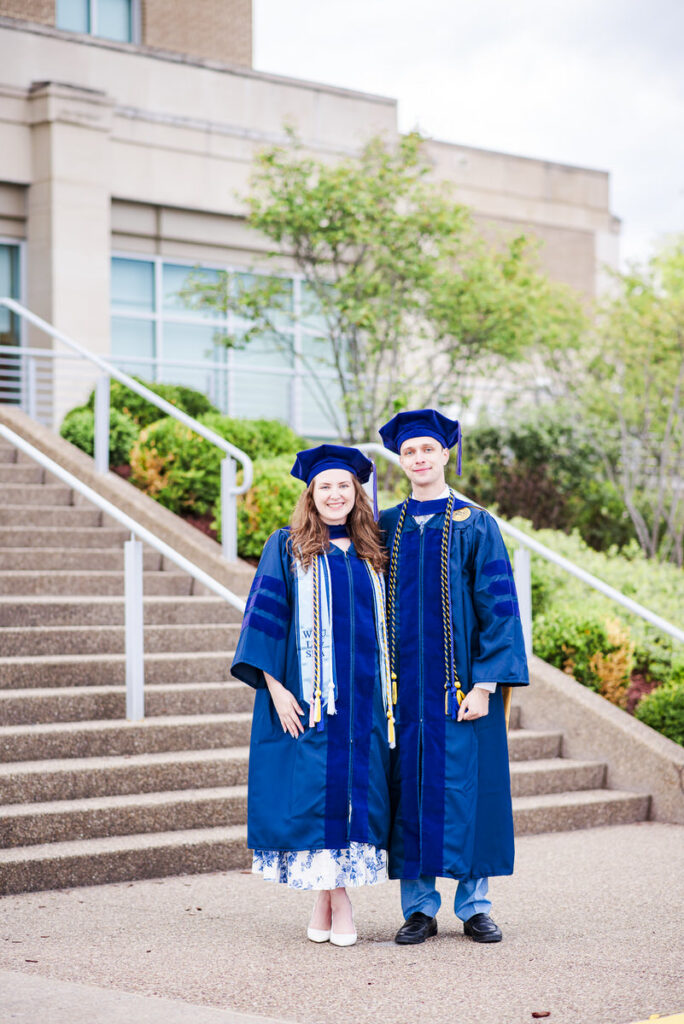 two graduates in blue gowns and caps pose on outdoor steps with a campus building behind them smiling for the photo