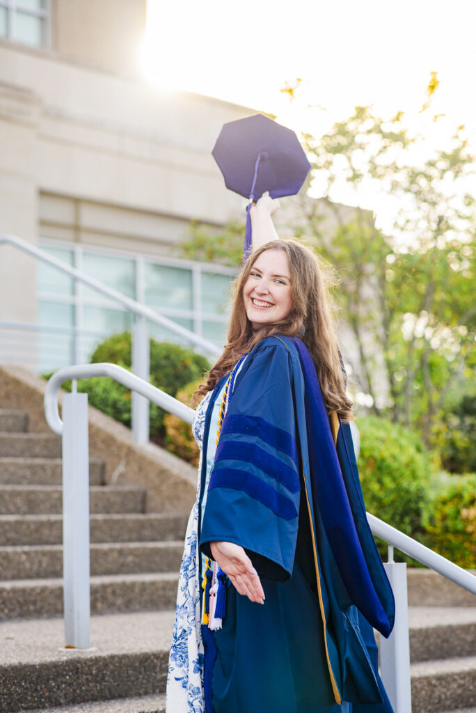 smiling graduate in a blue cap and gown raises her mortarboard outdoors on campus steps at sunset