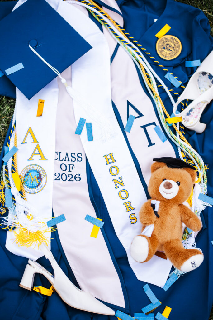graduation attire with blue gown white sashes reading'Class of 2026' and 'Honors', cords, a teddy bear, and a decorative high heel shoe on the grass.