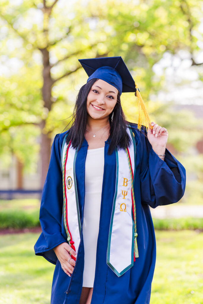 graduate in blue cap and gown outdoors holding a yellow tassel with white and greek letter stoles visible