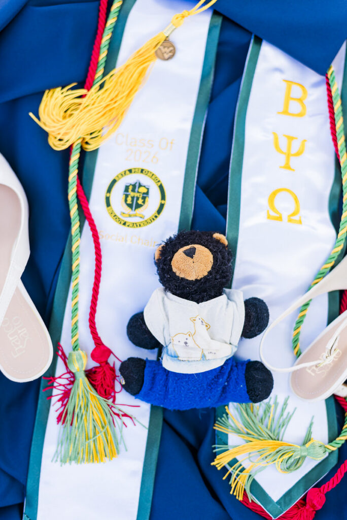 blue graduation gown with a white stole showing'Class of 2026,' plus colorful cords, tassels, and a small bear plush in a blue outfit.
