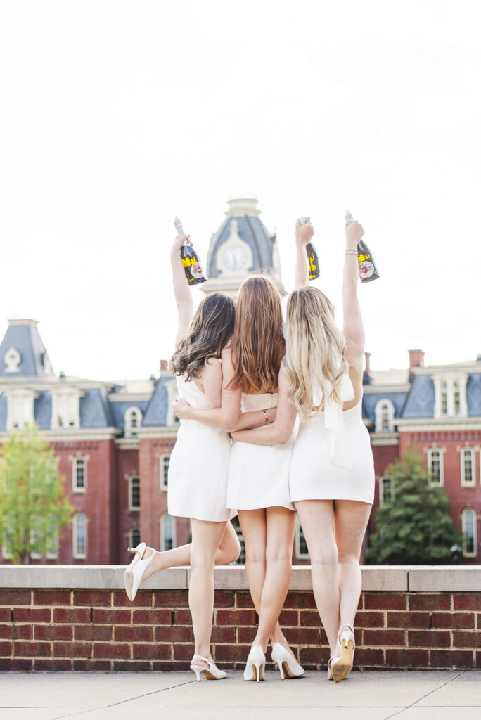 three women in white dresses stand with backs to the camera arms raised holding champagne bottles against a cityscape backdrop on a rooftop edge