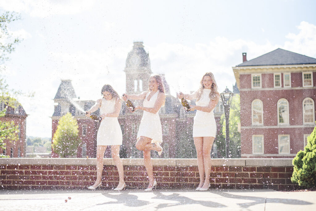 wvugradphotos286 carrie white photography three women in white dresses celebrate on a brick ledge spraying champagne as water droplets fly around with historic buildings in the background during wvu graduation