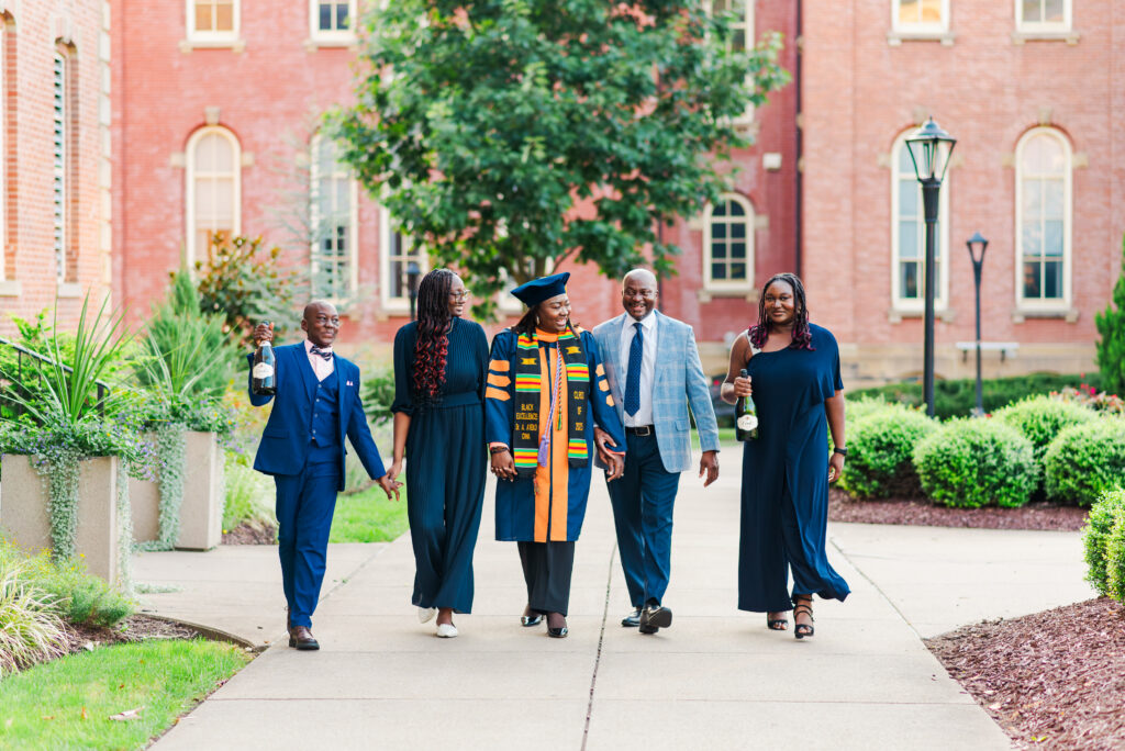 five graduates in blue gowns and formal attire walk arm in arm down a campus path holding champagne bottles and smiling