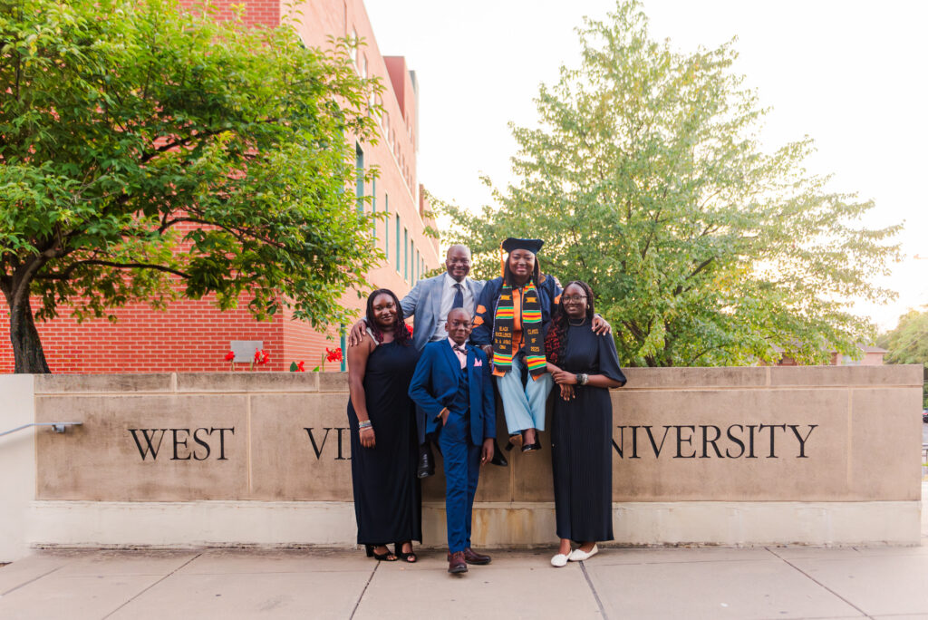 five graduates in caps and gowns posing on a stone wall that reads west university with trees and a brick building behind them