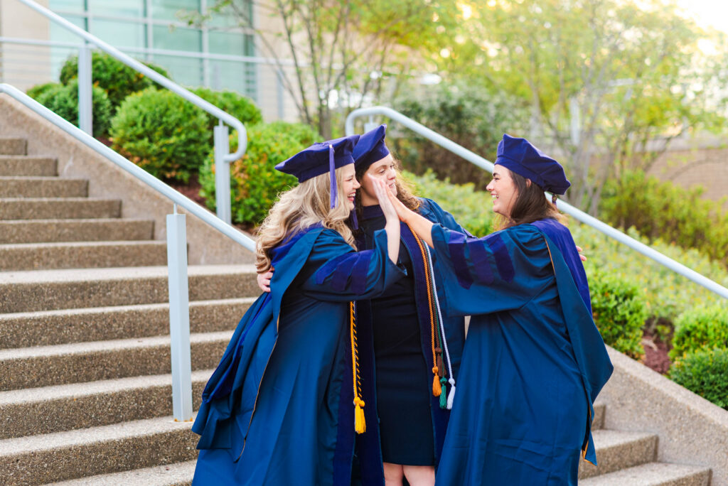 three graduates in blue gowns celebrate with a high five on campus stairs outdoors during wvu college of law grad photos