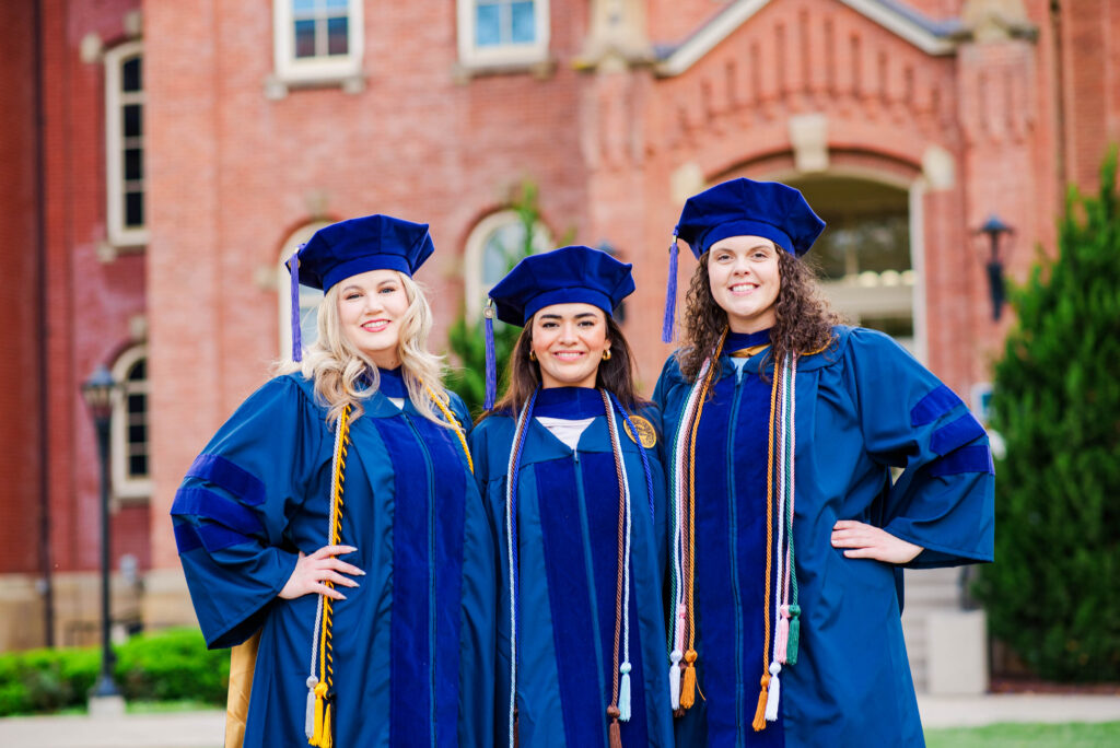 college of law wvu61 carrie white photography three graduates in blue gowns and caps posing outdoors in front of a brick campus building
