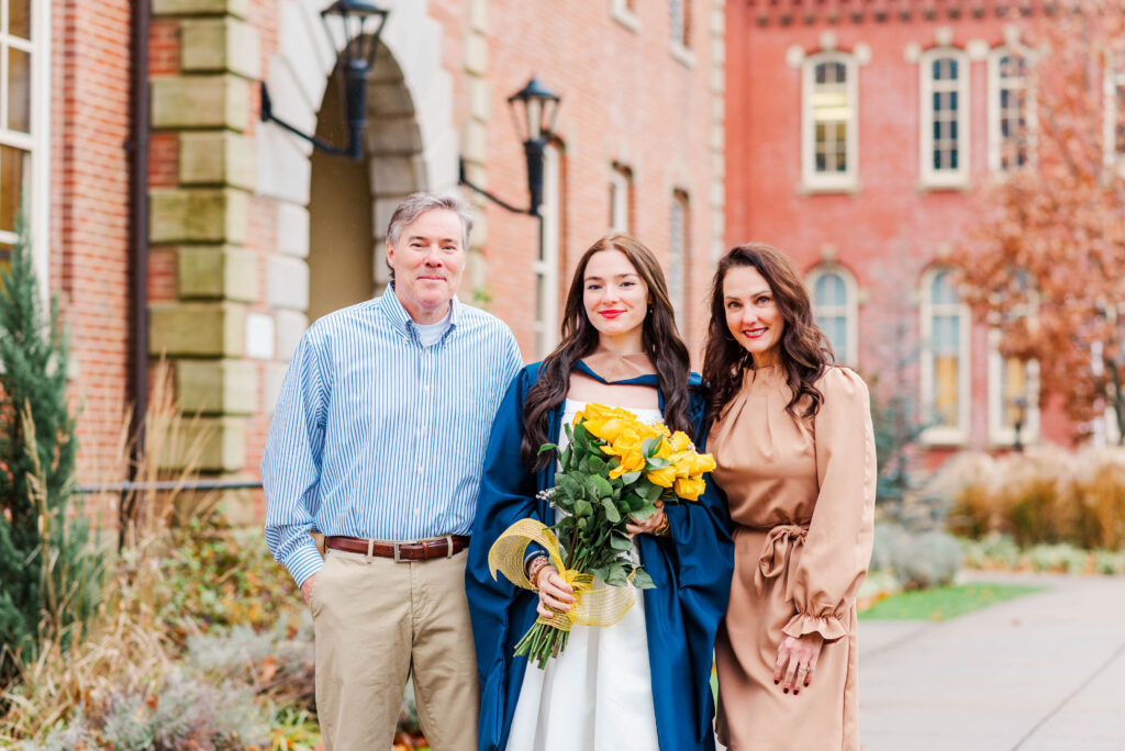 three people pose outdoors on a college campus after graduation the graduate in a blue gown holds a yellow bouquet while two friends stand beside her