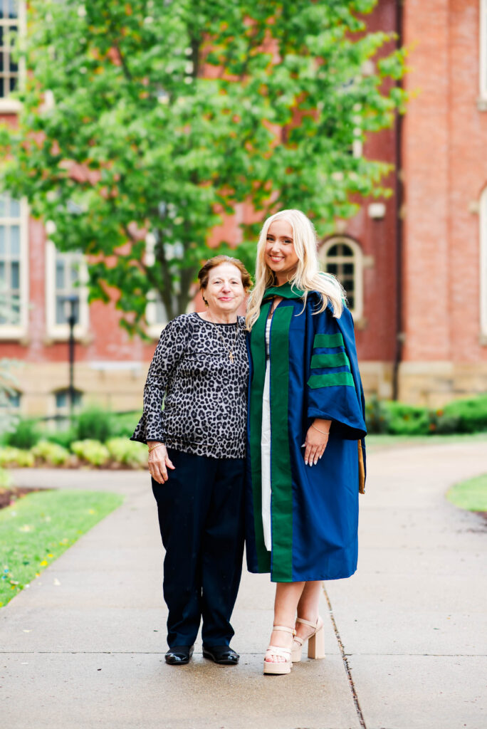 two women pose on a campus path a graduate in a blue gown with green trim beside an older woman in a leopard print blouse both smiling with a brick building and greenery in the background
