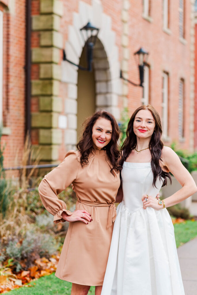 two women pose together outdoors wearing beige and white dresses smiling in front of a brick building