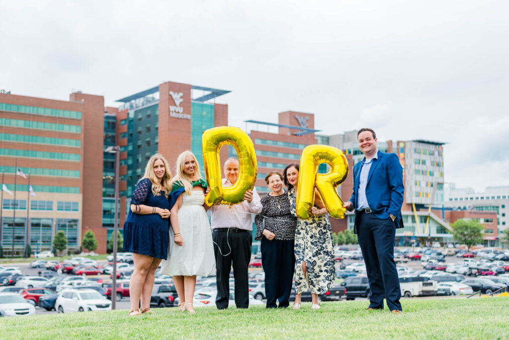 group of adults smiling and posing on a grassy area outside a modern brick and glass building with large gold balloons partially visible in the foreground