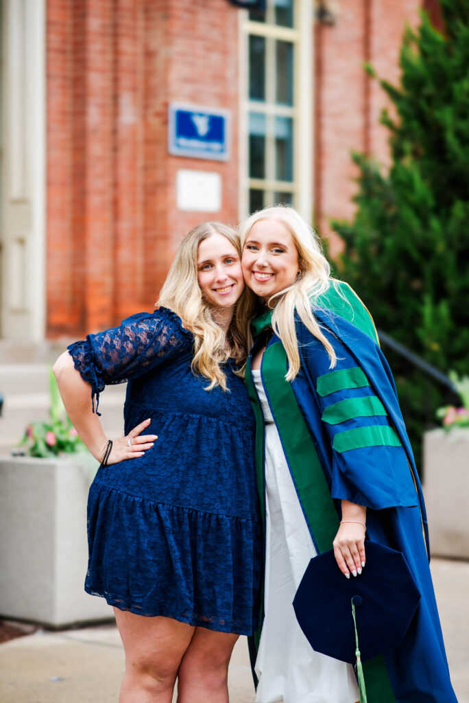 two young women in graduation attire smiling and hugging outdoors in front of a brick building