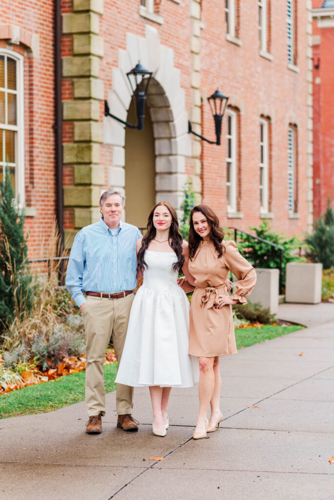 three adults posing on a sidewalk in front of a red brick building woman in a white dress stands center with a man on her left and a woman in a tan dress on her right