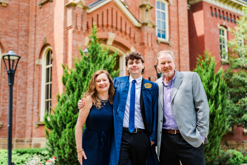 graduate in a blue gown stands with a smiling parents outside a red brick university building
