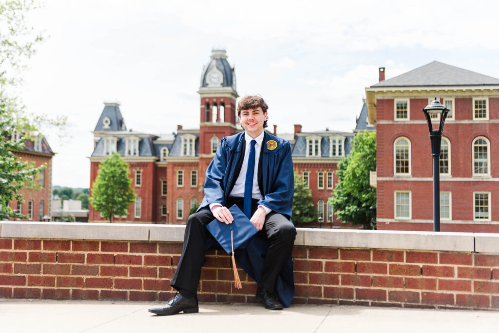 graduationphotoideasfor guys010 carrie white photography graduate in a blue gown and tie sits on a brick ledge with campus red brick buildings behind him and a clock tower in the background