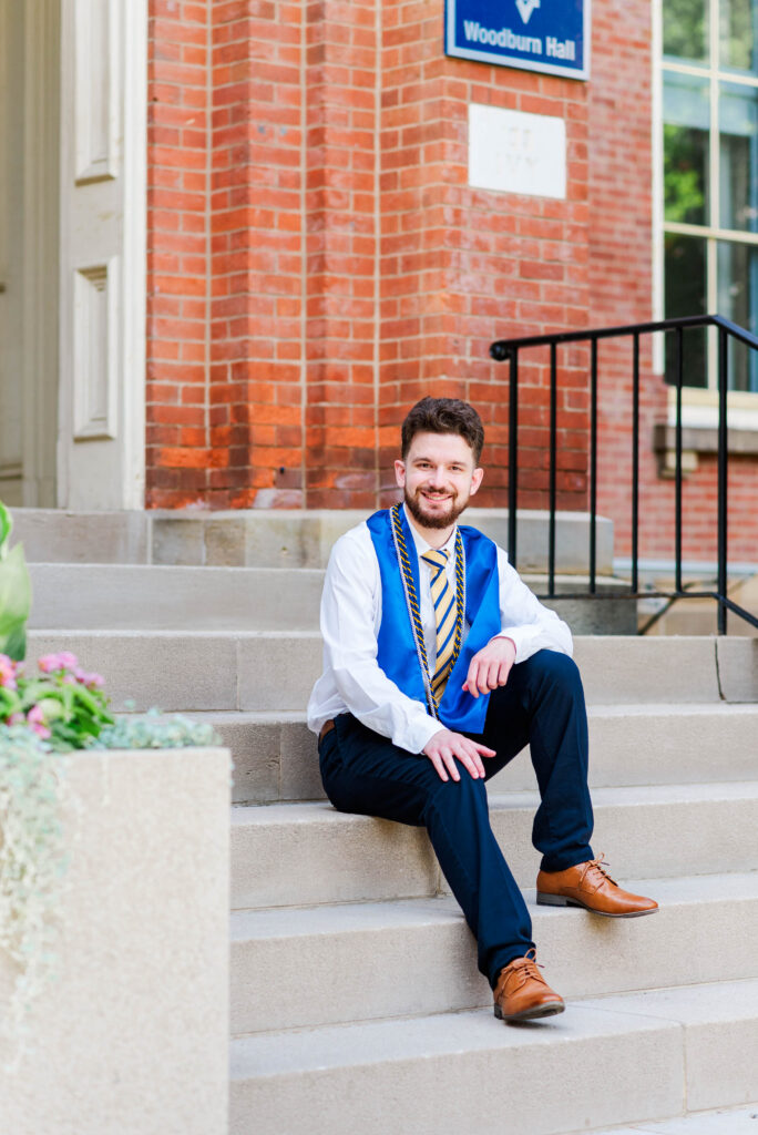 graduationphotoideasfor guys012 carrie white photography graduate student in a blue gown and gold stole sits on campus steps outside woodburn hall smiling at the camera