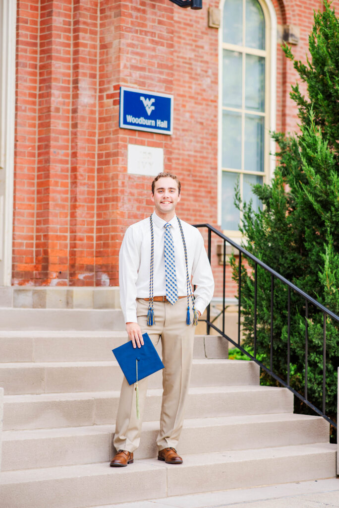 graduationphotoideasfor guys014 carrie white photography graduate standing on campus steps outside woodburn hall holding a blue diploma cover with cords around his neck