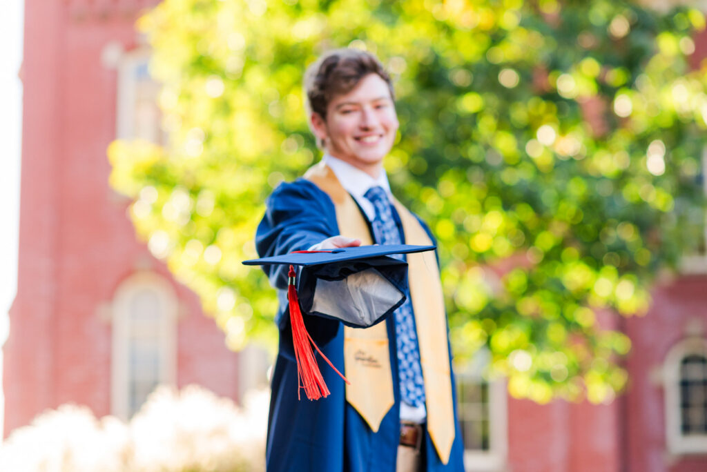 graduationphotoideasfor guys033 carrie white photography graduate in blue cap and gown throwing graduation cap toward camera outdoors greenery and brick building in background